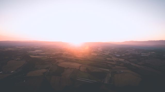 This picturesque scene captures an aerial view of a serene countryside at sunrise. The morning light creates magical hues over the horizon, providing a tranquil and peaceful atmosphere. The panoramic view of farmlands and fields can be used to depict concepts of serenity, tranquility, and the beauty of rural life. Ideal for websites, promotional materials, backgrounds, and inspirational posters.
