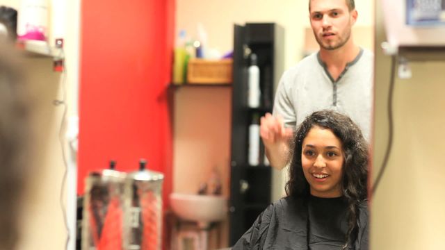 Scene captures young woman receiving haircut at a modern salon. Professional hair stylist behind focused on work while client sits in chair with satisfied expression. Useful for articles or promotions related to beauty, hairstyle trends, salon experiences, or professional hair care services.