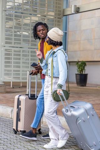 Diverse young travelers walking curbside pulling luggage and checking smartphone