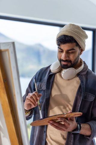 Indian male artist mixing paint on palette at easel wearing headphones and beanie