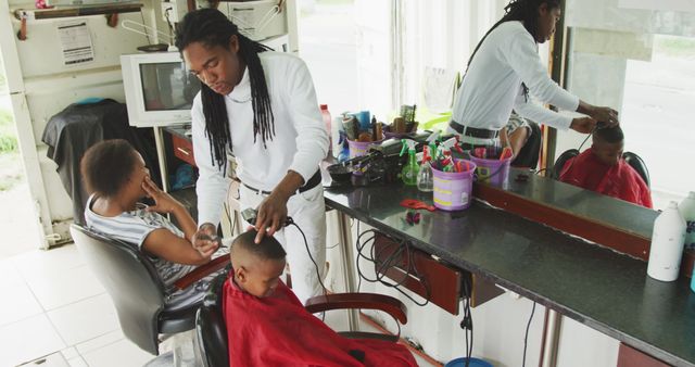Barber with dreads providing a haircut to a young boy wearing a red cape while a woman looks on beside them. Interior of afro caribbean barbershop, various grooming tools and products visible. Ideal for illustrating personal grooming and community-focused services.