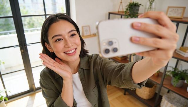Smiling asian woman taking selfie with smartphone in sunlit modern home interior lifestyle