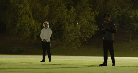 Diverse friends standing on floodlit golf fairway at night, sharing ball, walking together