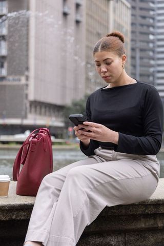 African american businesswoman checking smartphone by urban fountain with red tote and coffee