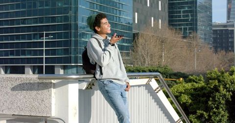 Young man leaning on railing holding smartphone on urban terrace with city skyline