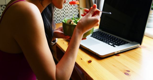 Woman working from home eating a healthy salad while using a laptop. Ideal for content on healthy living, remote work lifestyle, productivity tips, and modern nutrition. Could be used in blogs, articles, or advertisements related to work-life balance and wellness.