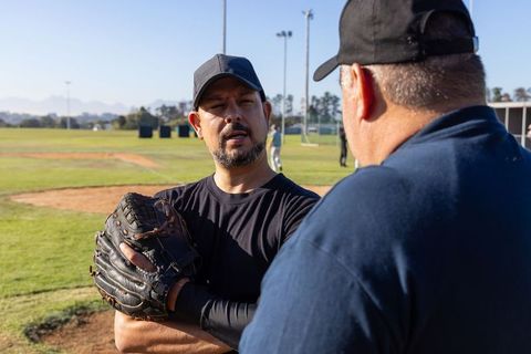 Coach giving pitching instruction to mature player holding glove on sunlit baseball field