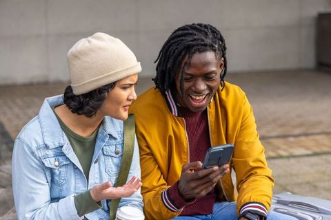 Multicultural friends laughing and sharing smartphone on urban bench, coffee and luggage
