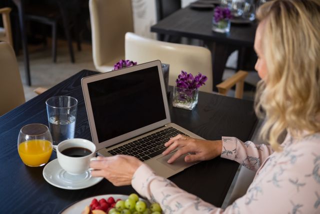 Businesswoman working on laptop in a restaurant while holding a coffee cup. Ideal for illustrating remote work, professional settings, and modern work environments. Suitable for articles on productivity, business lifestyle, and technology in everyday life.