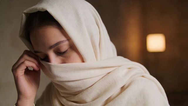 Woman in cream headscarf touching face and closing eyes with warm lamp glow