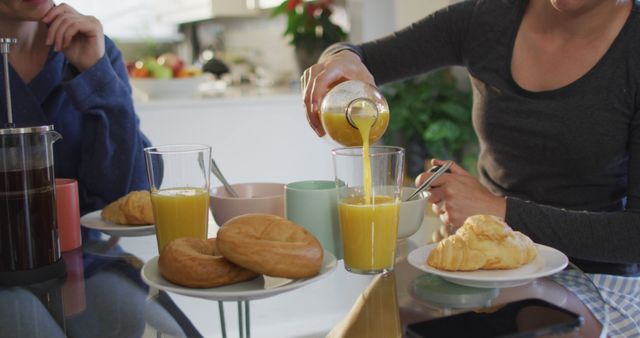 Two women enjoying breakfast together, with fresh croissants and glasses of orange juice on the table in a cozy kitchen. This image can be used for lifestyle blogs, food and drink marketing, or social media content promoting breakfast habits.