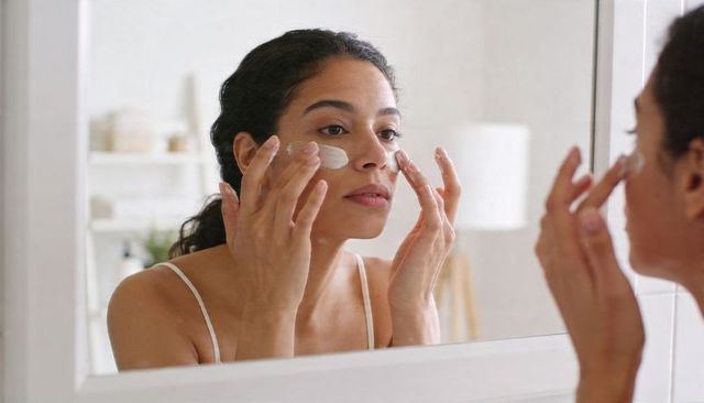 Applying moisturizer morning skincare routine woman rubbing cream on cheeks at mirror