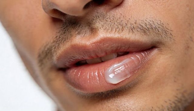 Close-up showing male lower face with glossy white balm droplet on lip, light stubble