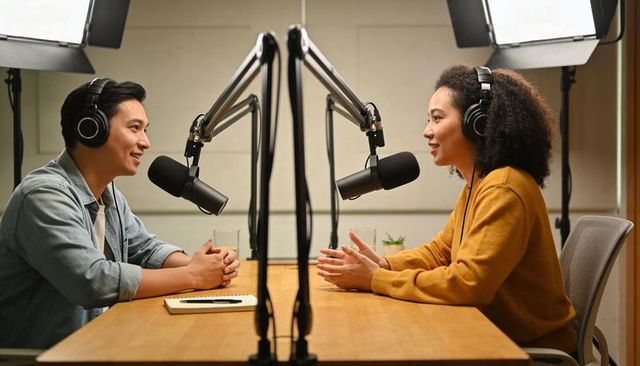 Two podcasters recording in modern studio wearing headphones and talking into microphones