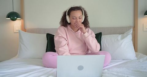 African american woman studying on bed in pink sweatsuit with headphones and laptop at home