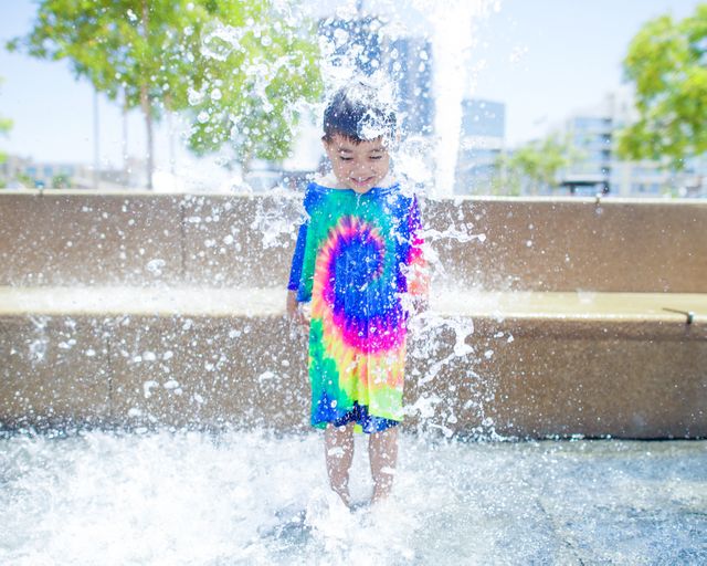 Child stands in colorful outfit enjoying refreshing water fountain on a sunny day in urban environment. Suitable for concepts of summer fun, childhood joy, outdoor activities, cooling off in heat, and family vacation. Great for promotional materials, blogs, and ads focusing on family-friendly occasions and summer recreation.