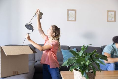 Young couple unpacking in living room lifting lamp while partner checking smartphone