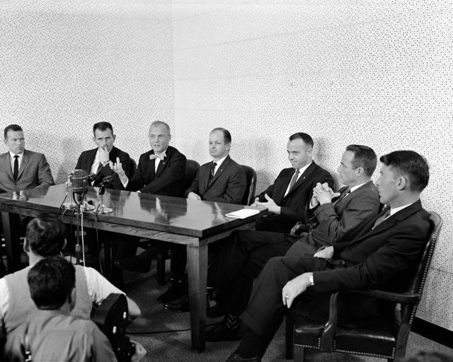 Mercury astronauts at a 1961 press conference discussing space missions with Col. John A. Powers. Astronauts seated around a table in formal suits while reporters listen attentively. Use for history of space exploration, significant moments in NASA history, or educational materials about early space missions.