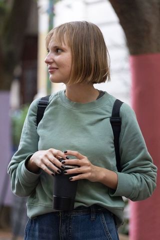 Young woman holding reusable travel mug, wearing sage sweatshirt and backpack on urban street