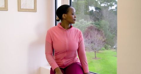 African American woman sitting on window ledge wearing pink activewear gazing at garden