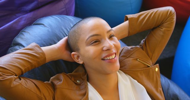 A young bald woman is seen smiling contentedly while leaning back on a couch. She wears a caramel brown leather jacket over a white top, exuding a casual and laid-back vibe. This can be used in lifestyle blogs, mental health articles, fashion features, or advertisements promoting joy and relaxation.