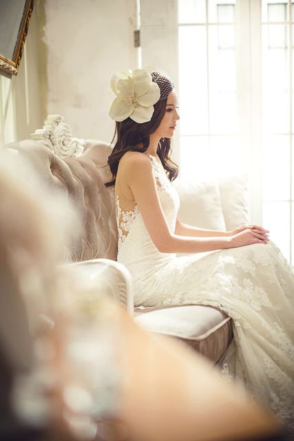 Bride wearing lace wedding dress sitting on vintage sofa with large flower in hair, bathed in sunlight. Perfect for wedding invitations, bridal fashion catalogs, romance novels, or wedding blogs highlighting bridal preparations and serene moments.