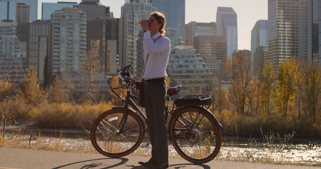 A person wearing casual attire stands next to a bicycle enjoying a coffee by a river in an urban park setting. The city skyline is visible in the background with buildings towering above. Autumn trees with colorful foliage add warmth to the scenery. Ideal for themes centered around outdoor activities, urban lifestyle, relaxation, and the blend of nature and city life.