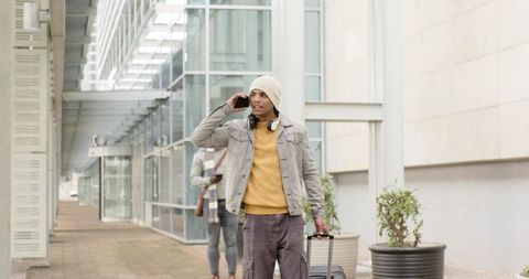 Young diverse men walking with luggage and using phones near modern glass building