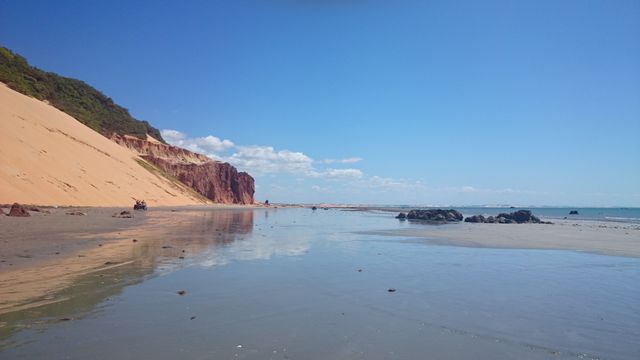 Idyllic scene of an empty beach featuring rolling sand dunes and red cliffs extending into the distance under a bright blue sky. Reflective wet sand leads to calm waters, ideal for showcasing natural beauty, promoting tourism, or adding scenery to creative projects. Perfect for travel brochures, environmental campaigns, or digital backgrounds.