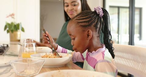 African american mother smiling while daughter enjoying pasta at kitchen counter