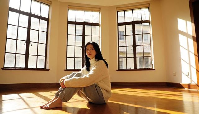 Sunlit minimalist interior with seated asian woman in cream sweater hugging knees on hardwood floor