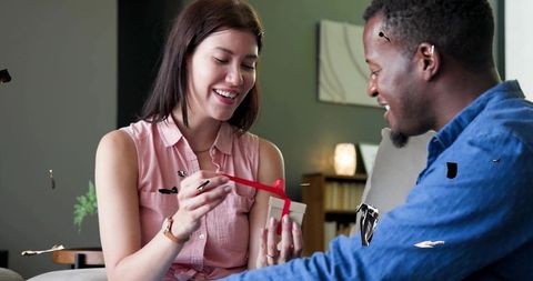 Couple opening gift on sofa at home smiling and unwrapping red ribbon present, cozy moment