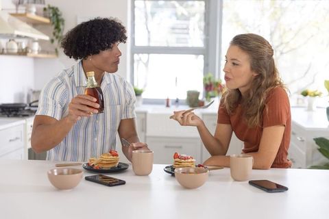 Diverse couple sharing pancakes and pouring syrup at bright modern kitchen island