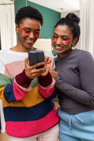 Diverse friends sharing eye patches skincare moment while laughing and checking smartphone