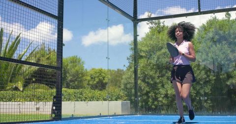 Athletic woman striding on blue padel court holding racket, outdoor sunny sport action