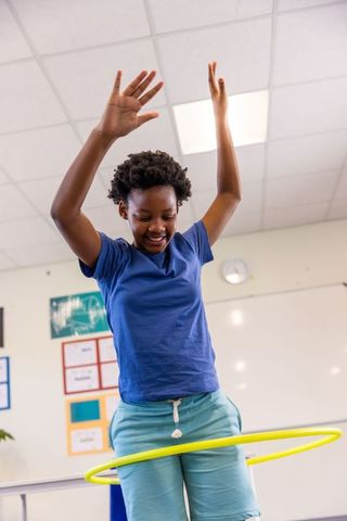 Joyful african american girl hula hooping in classroom raising arms during active play