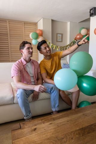 Two men celebrating birthday at home holding teal balloons and pointing on sofa