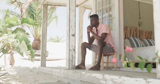 Man sitting in rustic cottage, contemplating while surrounded by tropical elements. Ideal for use in travel blogs, vacation retreats, mental health blogs, or serene lifestyle ads.