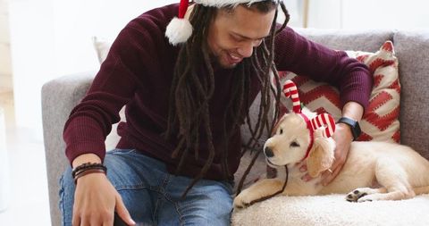 Man wearing santa hat petting golden retriever puppy wearing reindeer headband on cozy sofa