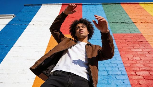 Young man raising arms against vibrant striped brick mural, urban fashion portrait