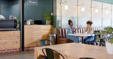 Two women chatting and sipping coffee at modern cafe booth with leather banquette and plants