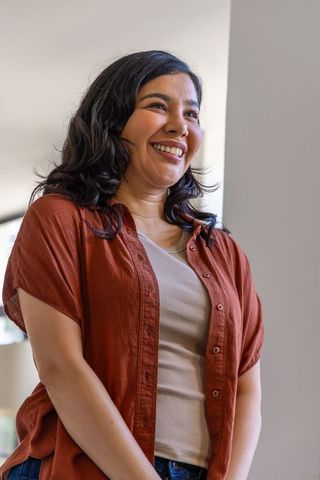 Smiling young woman standing in warm interior wearing rust shirt beige tank and denim