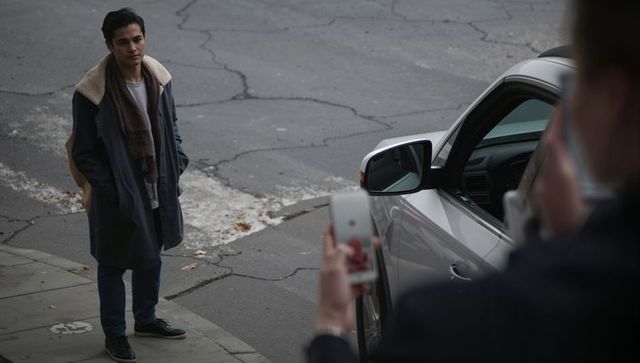 Young man standing and talking to driver beside silver sedan on cracked urban street