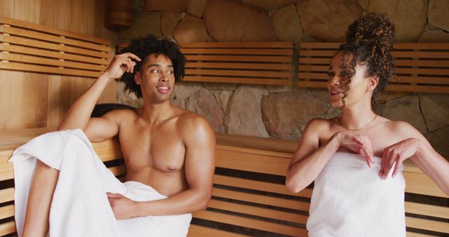 Image of relaxed happy diverse couple wearing towels sitting and talking in sauna room at health spa. Vacation, togetherness, relaxation, health and inclusivity concept.