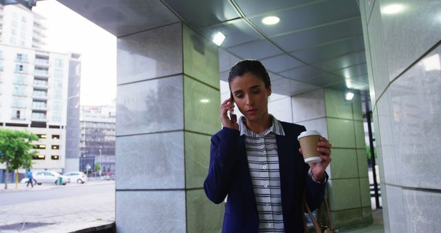 Businesswoman wearing formal attire and walking in urban environment, talking on the phone while holding coffee cup. Suitable for presentations, articles on business or professional lifestyle, commuting, or urban work environments.