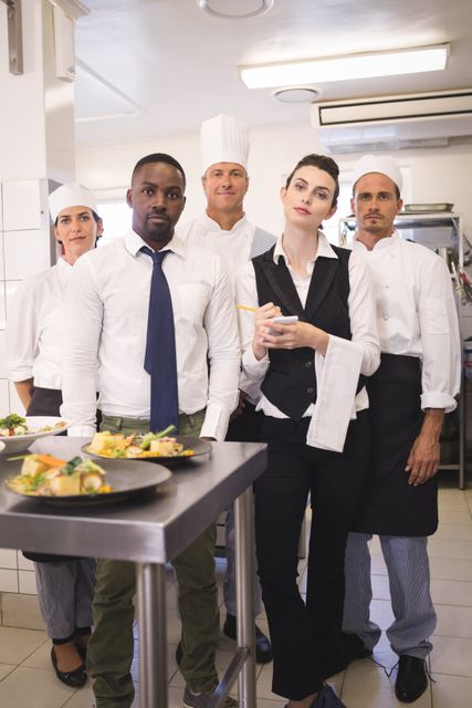 Restaurant manager standing confidently with a diverse team of chefs in a commercial kitchen. This image can be used for portraying teamwork, diversity in the workplace, and the professional environment within the culinary industry. Ideal for promotional materials, websites, and articles related to hospitality, restaurant management, and food service.