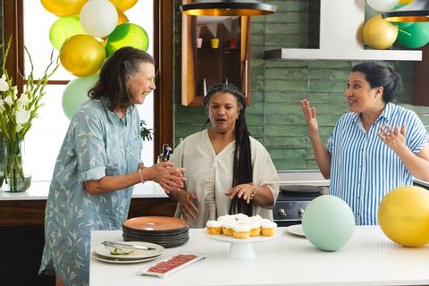 Diverse senior women celebrating at kitchen island with cupcakes and balloons