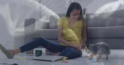 Smiling korean woman working remotely on floor at home petting small dog with laptop