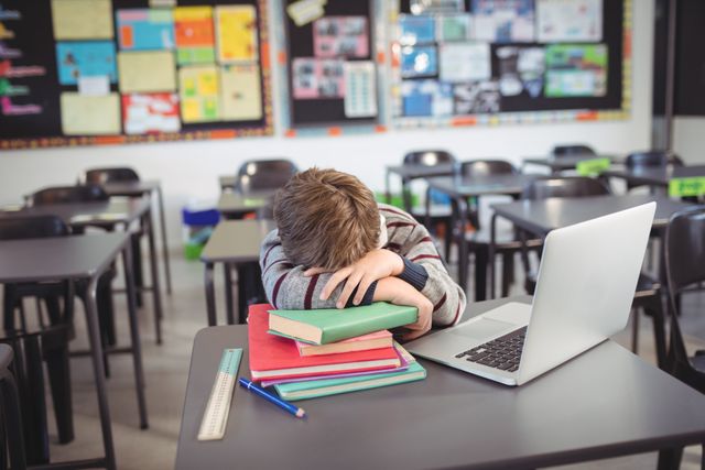 Tired schoolboy sleeping on stack of book by laptop in classroom