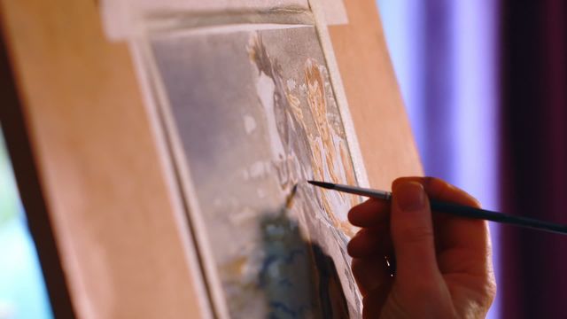 Female artist is painting a detailed scene on canvas at home. The video beautifully captures her hand holding a paintbrush as she works with close attention to detail. This can be used to represent creative hobbies, artistic process, or the concept of home studios. Suitable for use in articles about art, creativity, and home-based activities.